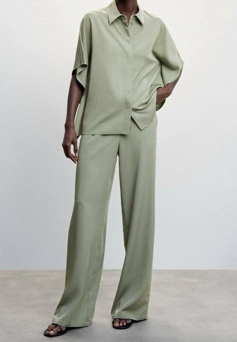 Personne portant une chemise ample à manches courtes vert clair avec boutons, un pantalon large assorti et des sandales noires, debout sur un sol gris.