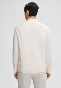 Maglione bianco, testurizzato, con un alto collo, maniche raglan e polsini a coste. La vista dal retro mostra un design liscio e aderente.