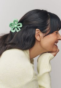 Pince à cheveux en plastique vert en forme de fleur avec un motif de pétales évidés. Placée sur des cheveux foncés et ondulés, portée avec un pull jaune clair.
