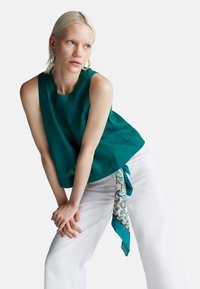 Tyrkysová lněná bezrukávová blůza s kulatým výstřihem, volným střihem a uvázanou dekorativní šálou z potištěné látky na bílých kalhotách.