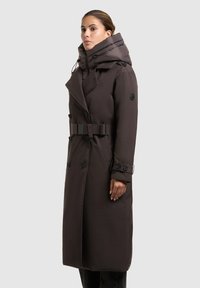 Donna che indossa un lungo cappotto marrone scuro con cintura, ampio colletto imbottito, dettagli con bottoni e zip sul davanti, su uno sfondo grigio uniforme.