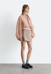 Helle rosa oversized Bluse mit Puffärmeln, kombiniert mit einem beige Minirock und einem weißen Gürtel, ergänzt durch schwarze Loafers und graue Socken.