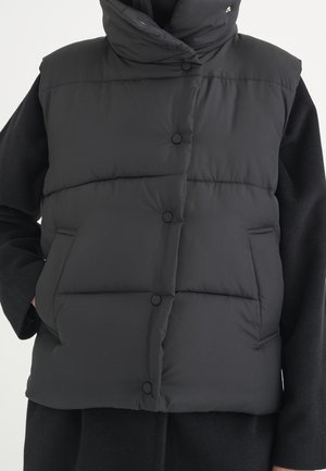 Czarna pikowana kamizelka na zatrzaski z bocznymi kieszeniami, noszona na czarnym płaszczu z długimi rękawami, ręce w kieszeniach, pokazany górny tułów.