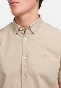 Beige overhemd met knopen gemaakt van geweven stof. Heeft een kraag, een voorflap en een klein geborduurd logo aan de linkerzijde.