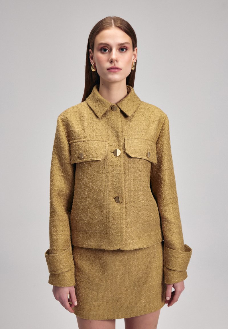 Giacca gialla in tessuto con tasche frontali, chiusura con bottoni e polsini larghi. La gonna abbina la giacca per materiale e colore. Design minimalista.