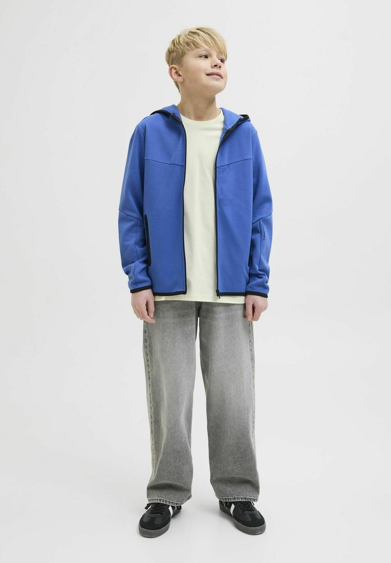 Chaqueta de forro polar azul con cremallera, con ribetes negros en contraste y dos bolsillos frontales, llevada sobre una camiseta de color amarillo claro y pantalones de pierna ancha grises.