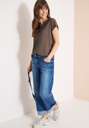 Uśmiechnięta kobieta w brązowej bluzce z krótkim rękawem, podwiniętych niebieskich dżinsach i srebrnych trampkach, trzymająca czarny pasek, stojąca na tle jasnej ściany.