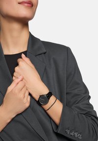 Schwarze Armbanduhr mit glattem Armband, rundem Ziffernblatt und minimalistischem Design. Tragen mit einem grauen Blazer mit ausgeprägten Revers und Knopfdetails.