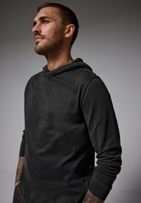 Svart hoodie med en mjuk textur, med rundad halsringning, långa ärmar och en avslappnad passform. Synlig sömnad och ribbade detaljer.