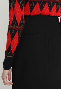 Personne portant une jupe noire et un pull à manches longues à motif argyle rouge et noir avec des coutures dorées, bras détendu sur le côté.