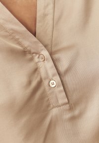 Ljus beige skjorta med krage i mjuk textur, prydd med små runda knappar och en subtil glans. Tyget verkar slätt och lätt.