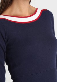 Navy blue long-sleeve top featuring a boat neckline with red and white stripes. The fabric has a ribbed texture and fitted design.