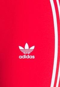 Červené sportovní legíny se bílými bočními pruhy a bílým logem Adidas. Materiál má hladkou, pružnou texturu.