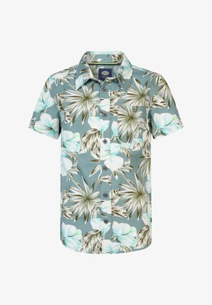 Camisa de flores de manga corta en azul verdoso y blanco, jeans azules y zapatillas blancas. La camisa presenta grandes patrones de hojas y un cierre de botones en la parte delantera.