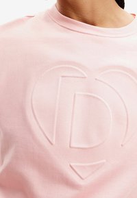 Hellrosa Sweatshirt mit einem geprägten Herz- und Buchstabenlogo, gefertigt aus weichem Baumwollstoff mit geripptem Rundhalsausschnitt.