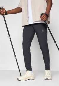 Mørkegrå treningbukser med smal passform og side lommer, paret med beige sneakers og beige skjorte, holder på turstaver.