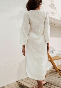 Bílé lněné šaty s dlouhými rukávy, kulatým výstřihem a přímým střihem. Mají centrální šev na zádech a délku ke kolenům.