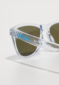 Przezroczyste okulary przeciwsłoneczne w kształcie kociego oka z ciemnymi soczewkami. Na zauszniku widnieje niebieska marka "OAKLEY". Teksturowane oprawki zwiększają chwyt.