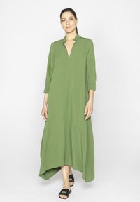 Zielona sukienka z długim rękawem, dekoltem w kształcie V i asymetrycznym hemem, uszyta z miękkiego materiału. Nosi się z czarnymi sandałami. Gładka faktura, minimalistyczny design.