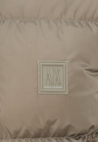Beige puffer jacket with a smooth, glossy finish. Features a square Armani Exchange logo patch in the center with embossed lettering.