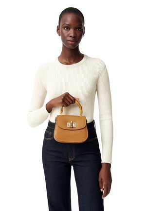 Vrouw in een witte geribbelde trui en donkere spijkerbroek, die een kleine beige handtas met bamboe handvat en gouden sluiting vasthoudt.