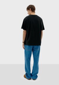 Schwarzes T-Shirt mit kurzen Ärmeln und rundem Ausschnitt, kombiniert mit lockeren blauen Jeans und braunen Schuhen, von hinten betrachtet.