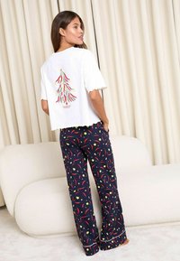Femeie îmbrăcată cu un tricou alb cu designul unui brad de Crăciun din ardei iute și pantaloni de pijama bleumarin cu modele de ardei iute roșu și stele galbene.