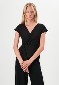 Haut noir à manches courtes, décolleté en V et détail frontal torsadé. Texture lisse et design ajusté. Aucune quincaillerie ou motif visible.