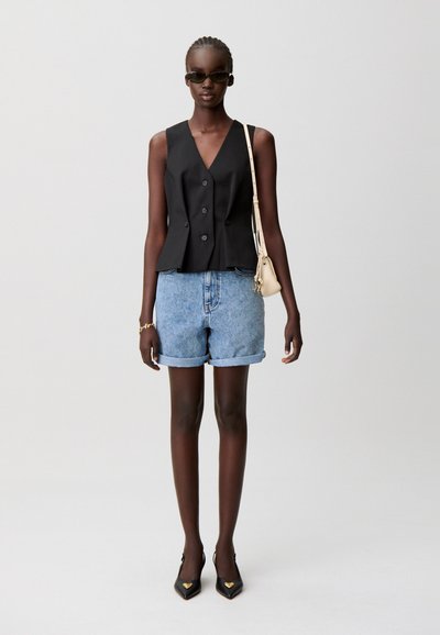 Blazer nero senza maniche con cinque bottoni, abbinato a pantaloncini di denim risvoltati e tacchi neri appuntiti con dettagli a forma di cuore.