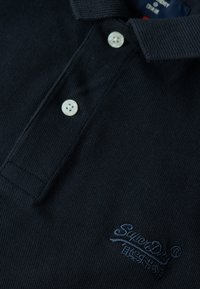 Marineblauwe polo gemaakt van een textuurstof, met een klassieke kraag, drie witte knopen en een geborduurd logo op de borst.