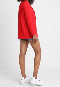 Personne portant un blazer rouge, un short en jean et des baskets blanches FILA, se tenant de profil contre un fond blanc uni.