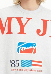 Grå sweatshirt med stor röd text, en logotyp i blått och rött, samt tryckta detaljer som inkluderar ett år och en amerikansk flagga.