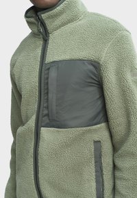Fleece bunda v svetlozelenej farbe s tmavozeleným panelom na hrudi, s vysokým golierom, zipovým zapínaním a dvoma bočnými vreckami.