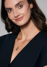 Złota naszyjnik z zawieszką, która posiada niebieskie i czarne kamienie, noszona na ciemnej odzieży, eksponująca delikatny łańcuszek i szczegóły.