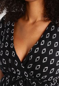 Robe noire cintrée avec un motif en losanges blancs, fabriquée en tissu doux, dotée d'un décolleté en V profond et de détails froncés à la taille.