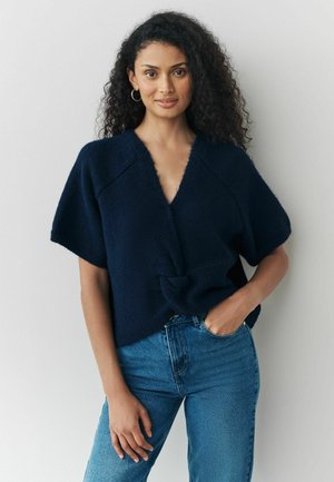 Kvinde med krøllet hår iført marineblå kortærmet slå-om sweater og blå jeans, står med den ene hånd i lommen mod en ensfarvet baggrund.