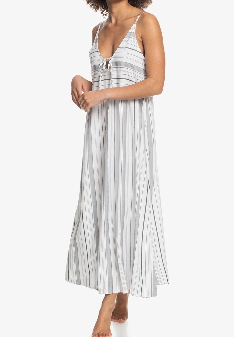 Femme portant une robe blanche sans manches, longue jusqu’aux chevilles, avec de fines rayures noires verticales et un détail noué à l’avant au niveau de la poitrine.