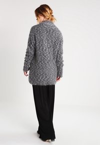 Šedý pletený cardigan s volným střihem, texturovaným vzorem, dlouhými rukávy a zaobleným lemem. Nositý přes černé kalhoty.