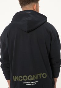 Felpa navy con grande scritta verde "INCognito" sul retro. Caratterizzata da cappuccio con cordino e vestibilità ampia. Texture liscia, elementi di design minimal.
