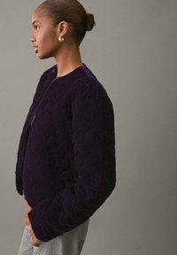 Giacca trapuntata viola con scollo rotondo, superficie texturizzata e maniche lunghe, abbinata a pantaloni in denim chiaro e orecchini semplici.
