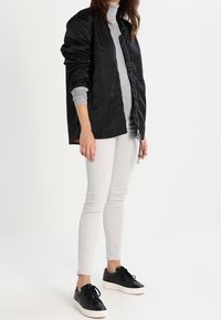 Schwarze Bomberjacke mit Reißverschluss, darunter ein grauer Rollkragenpullover, kombiniert mit hellgrauen Jeans und schwarzen Sneakers mit weißer Sohle.
