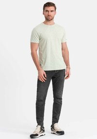 Jasnozielona koszulka z krótkim rękawem w mały kropeczkowy wzór, zestawiona z ciemnoszarymi dopasowanymi dżinsami i czarno-beżowymi butami sportowymi.