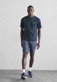 Marinefarbendes Baumwoll-T-Shirt mit weißem Logo, graue Shorts und marinefarbene Sportschuhe mit weißen Akzenten. Das Modell steht vor einem hellgrauen Hintergrund.