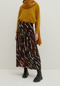 Pull en tricot jaune avec une capuche oversized, associé à une longue jupe plissée présentant un motif tie-dye marron, noir et blanc, ainsi que des bottes noires à lacets.