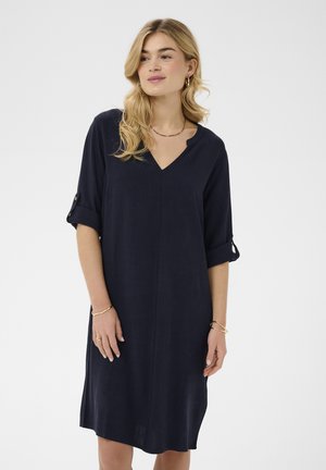 Szőke hajú nő, sötétkék, térdig érő, V-nyakú ruhát visel, felhajtott ujjakkal és arany ékszerekkel, fehér háttér előtt áll.