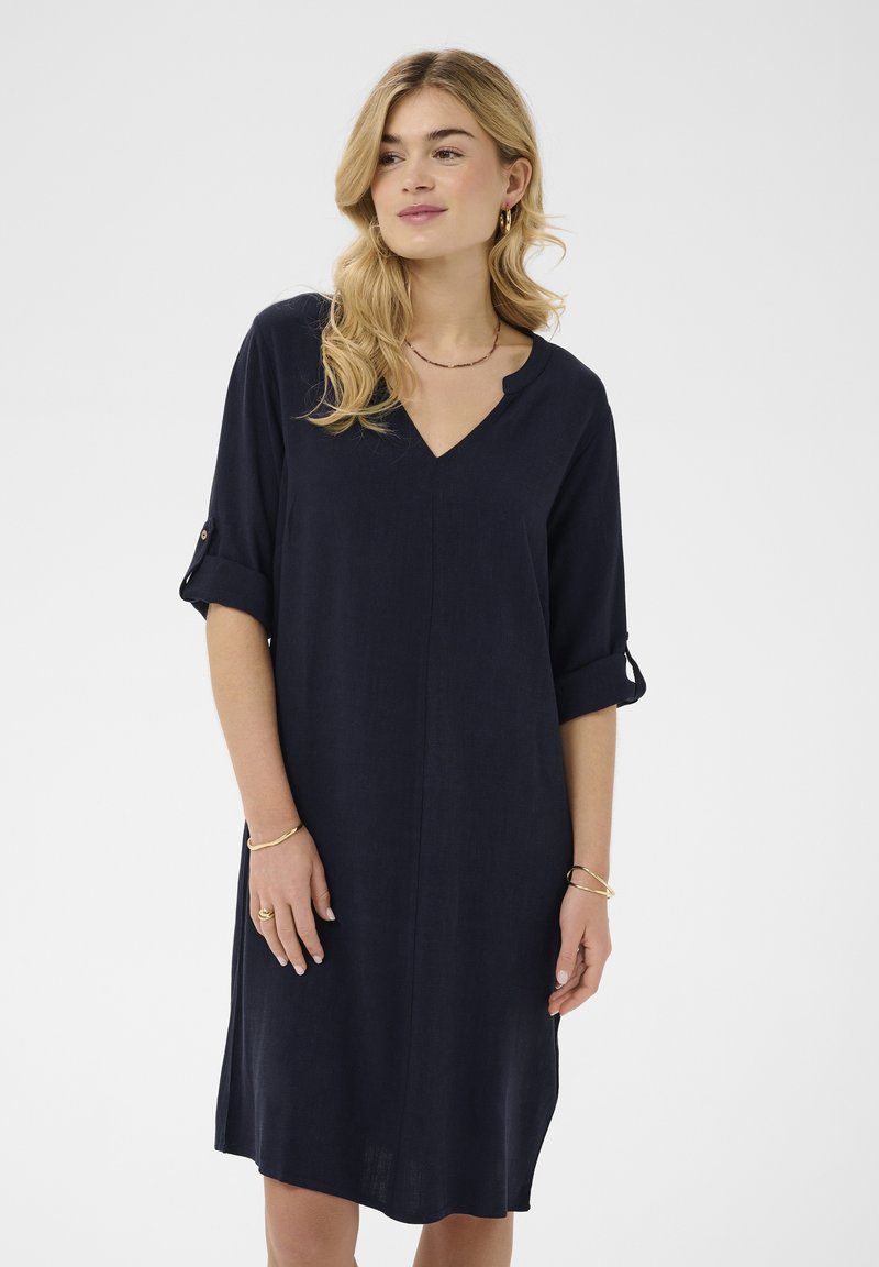 Femeie cu părul blond, purtând o rochie bleumarin, cu decolteu în V și mâneci suflecate până la genunchi, accesorizată cu bijuterii aurii, stând în fața unui fundal alb.