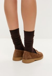 Mocasines de ante marrón con suela de goma, con un diseño sencillo y un acento plateado. Combinados con calcetines de canalé marrón oscuro.