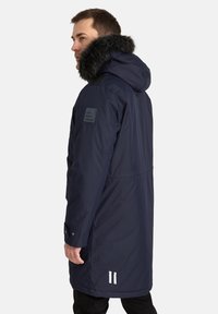 Parka blu navy con cappuccio foderato in pelliccia sintetica, spallina, tasche laterali e strisce bianche all'orlo; materiale impermeabile, design di lunghezza media.
