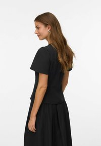 Czarna bluzka z krótkim rękawem o dopasowanym kroju, gładkiej fakturze i wyraźnie zarysowanych ramionach, w połączeniu z czarną spódnicą, wyeksponowana z boku.