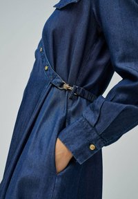 Vestido de mezclilla en azul oscuro con cinturón en la cintura, detalles de botones dorados, mangas largas y bolsillos laterales. Textura suave con un ajuste entallado.